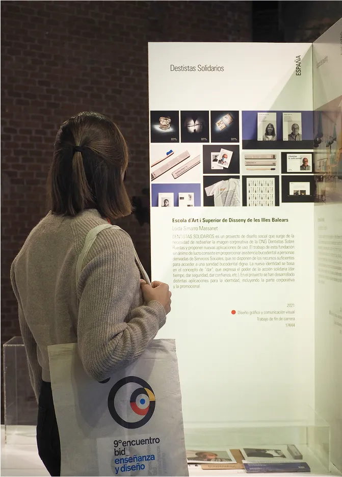Loida Simarro carrying a "9º Encuentro Bienal Iberoamericana de Diseño - Enseñanza y Diseño" tote bag while observing the "Dentistas Solidarios" project exposition.
