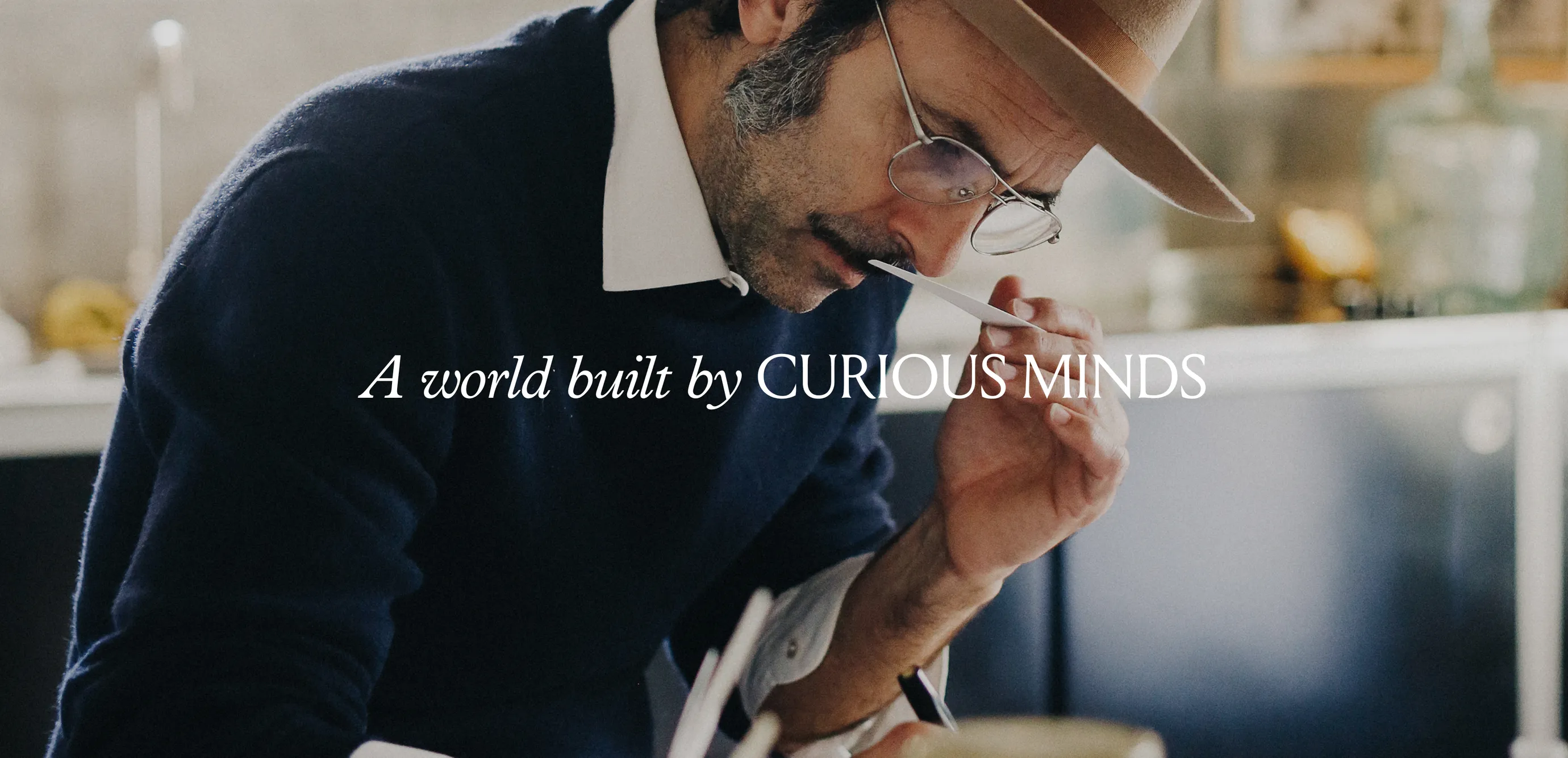 Man with glasses and hat in a blue sweater smelling a fragance while taking notes