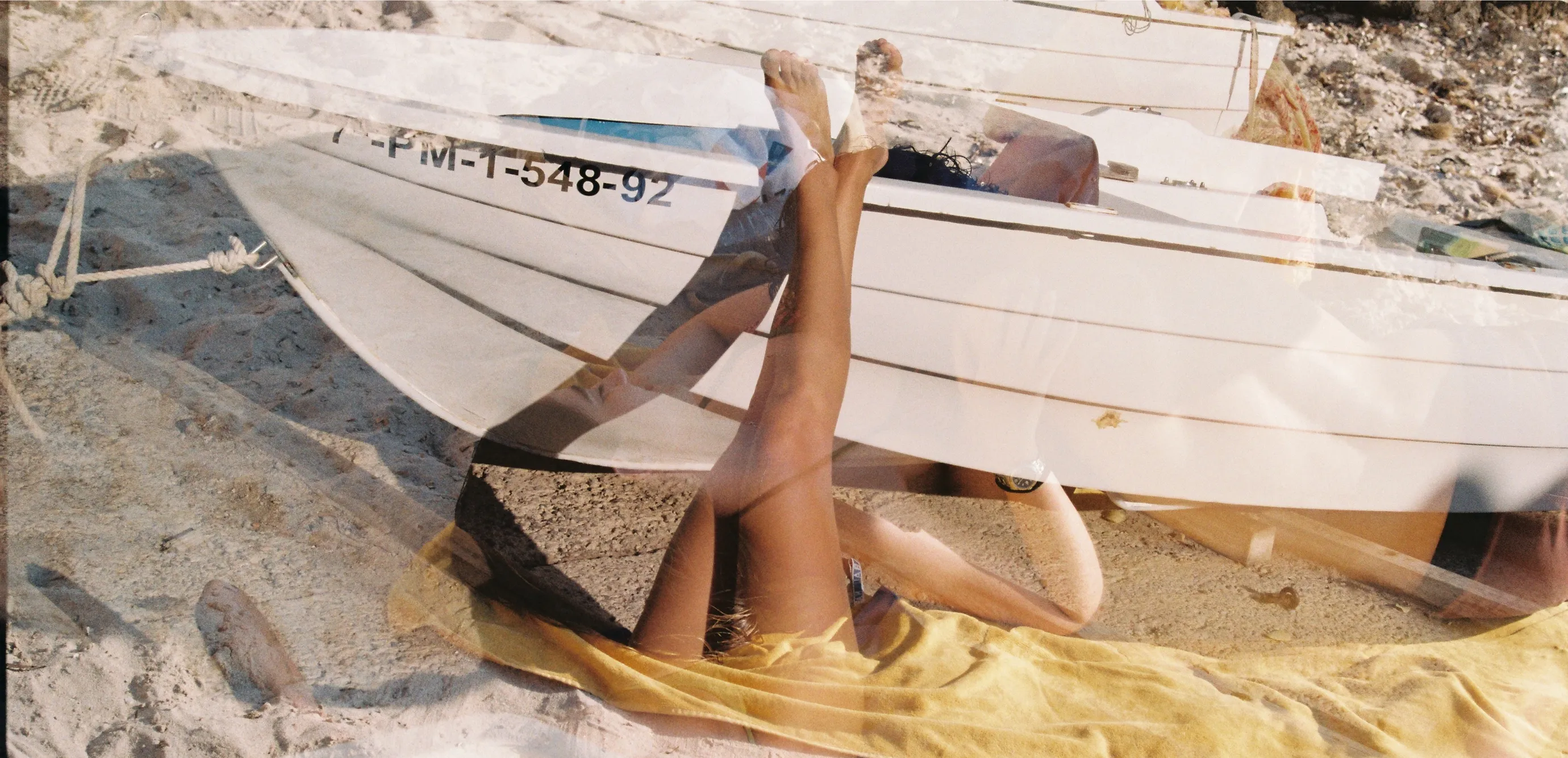 Double exposition photo, the first one being a white boat and legs and the second one a person laying down and reading on the beach.